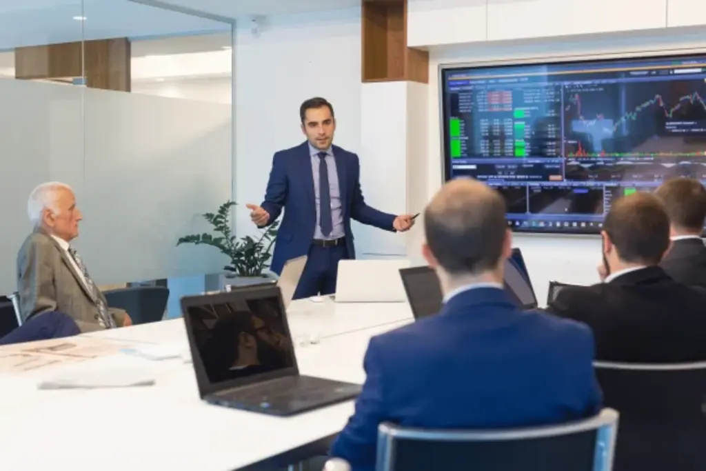 Corporate Training Courses- A presenter in a blue suit stands at the front of a modern conference room addressing a group of professionals seated around a table.