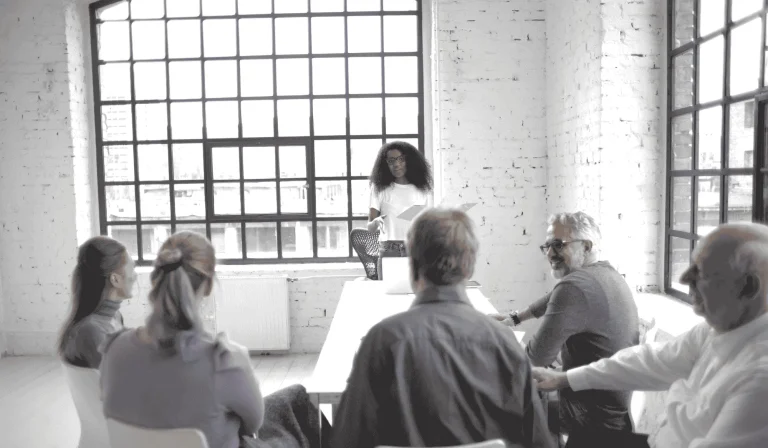 Online Leadership Training Courses - A woman stands at the front of a bright room presenting to a seated group of people gathered around a long table.
